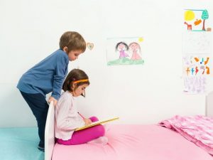 Girl sitting in bed drawing and brother watching over her shoulder